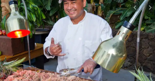 Chef prepares food under heat lamps, smiling and making a hand gesture near a buffet table.