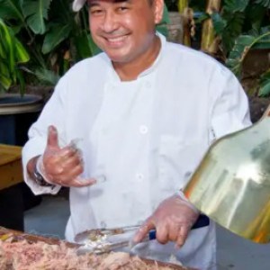 Chef prepares food under heat lamps, smiling and making a hand gesture near a buffet table.