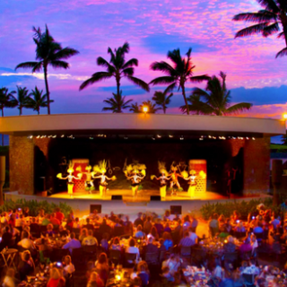 Outdoor stage with dancers and palm trees at sunset, audience seated at tables.