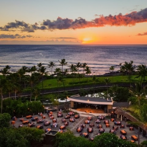 Sunset over ocean with outdoor dining area and palm trees in foreground.