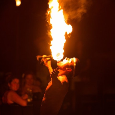 Performer breathes fire at a night event, with audience watching in the background.